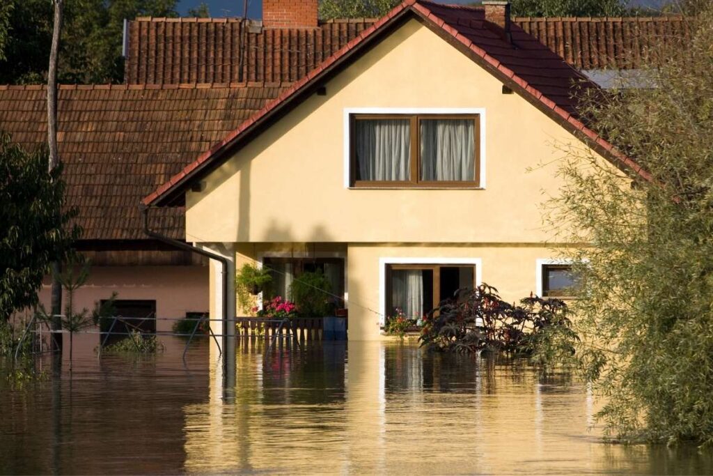 Maison beige partiellement submergée par l'eau, entourée de végétation, avec des fenêtres à volets fermés et un toit en tuiles rouges.