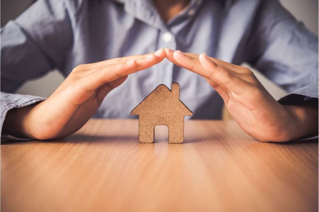 Deux mains entourent une petite maison en bois sur une table en bois clair.