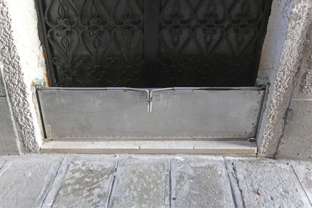 Une barrière anti-inondation métallique est installée devant une porte noire ornée de motifs décoratifs, sur un sol pavé gris.