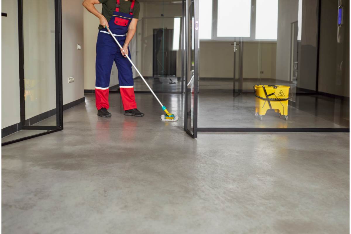 Un homme en salopette nettoie un sol gris avec une serpillière, à côté d'un seau jaune, dans une maison après une inondation.